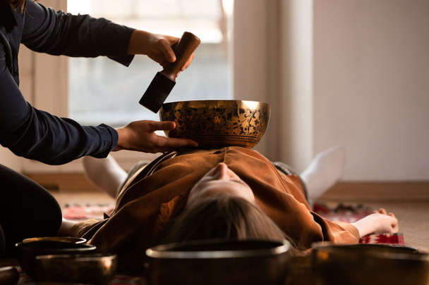 Woman making relaxing massage, meditation, sound therapy with tibetian singing bowls. Stress relief. Selective focus on bowl. Bottom view. - Photo, Image