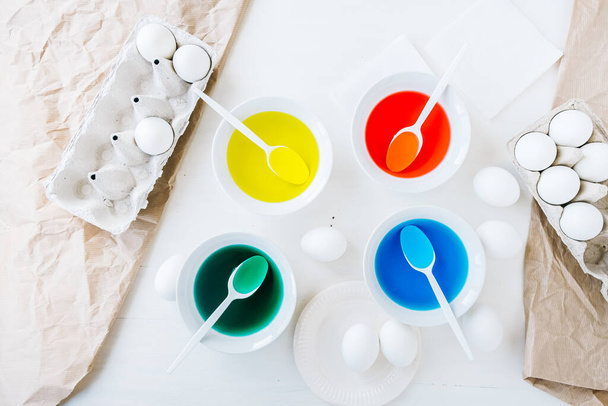Preparing for dyeing Easter eggs with liquid food coloring. Different colors in different bowls, egds on white table and craft paper background - Photo, Image