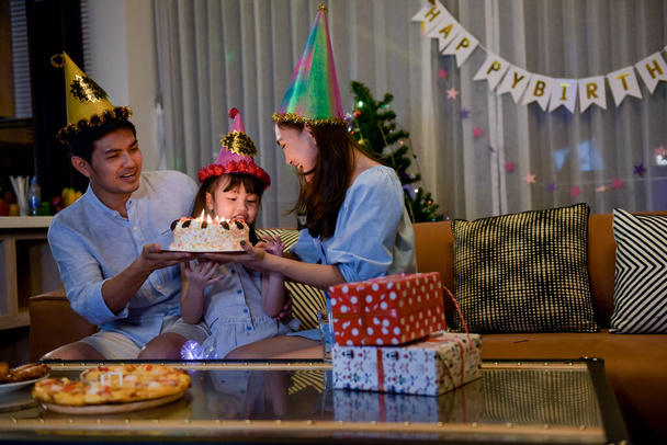 Ευτυχισμένη οικογένεια απολαμβάνει με την κόρη γιορτάζει τα γενέθλια μαζί, ενώ τούρτα γενεθλίων φυσάει στο σαλόνι στο σπίτι, που είναι όμορφα διακοσμημένα και κουτί δώρου. - Φωτογραφία, εικόνα