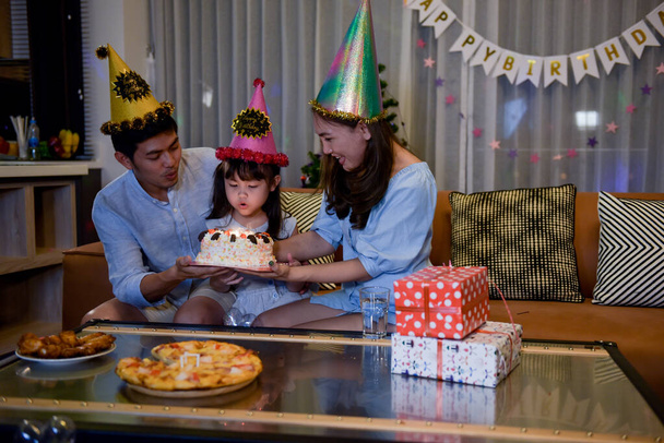 La familia feliz disfruta con la hija celebrando cumpleaños juntos mientras que el pastel de cumpleaños que sopla en la sala de estar en casa, que están bellamente decoradas y caja de regalo. - Foto, imagen