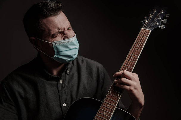um homem com uma máscara médica num fundo escuro, canta com uma guitarra acústica antiga. Retrato de estúdio - Foto, Imagem