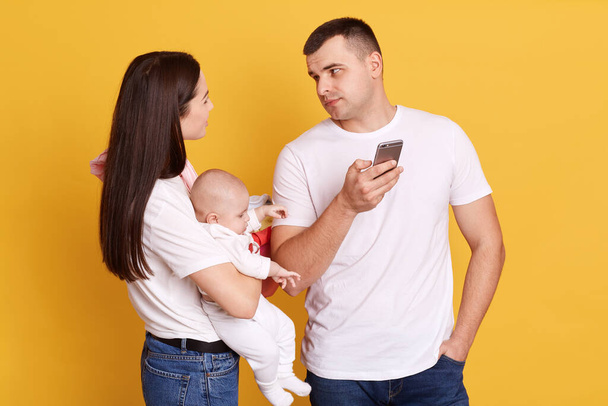 Young attractive mother with newborn daughter in hands looking at her husband and makes complaint because father does not help with house hold chores and child, posing isolated over yellow background. - Foto, afbeelding
