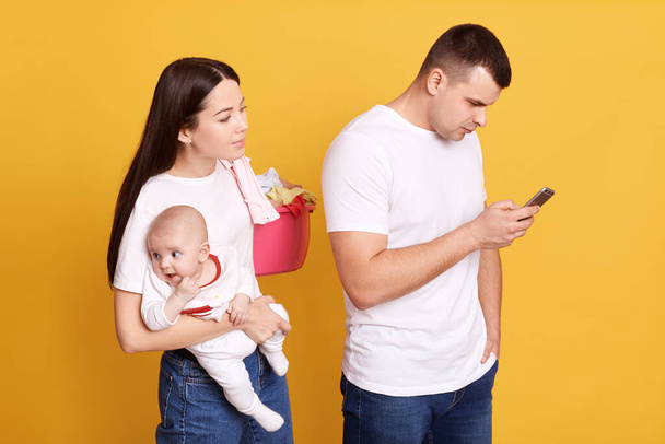 Jealous wife spying phone of her partner posing behind him with newborn child and basin with linen while male checking his social network. Couple posing isolated over yellow background. - 写真・画像