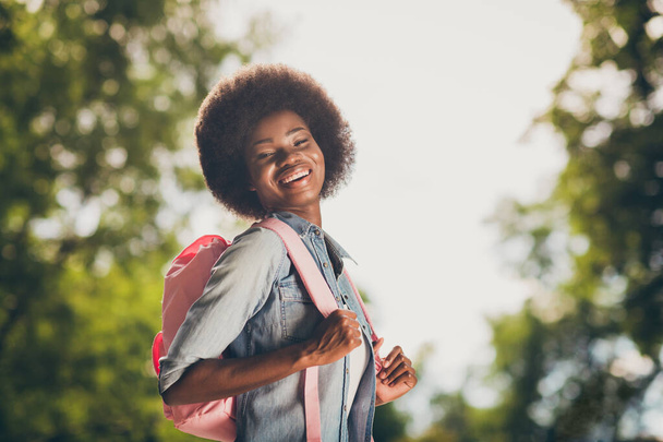 Side profile photo of black skinned pretty girl with curly hair laughing happily keeping backpack smiling in green city park - 写真・画像