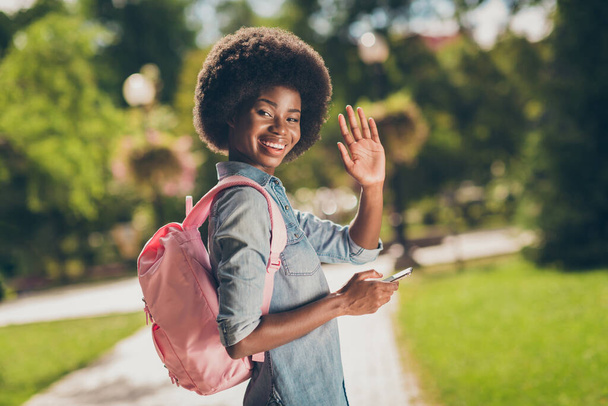 Side profile photo of black skinned female student keeping smartphone walking in sunny park waving with hand looking back smiling - Fotografie, Obrázek