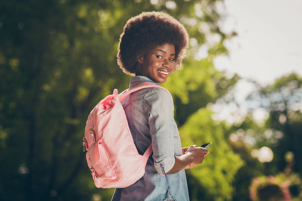 Rear view photo portrait of black skinned pretty girl keeping mobile phone walking in sunny park wearing pink backpack smiling - Photo, image