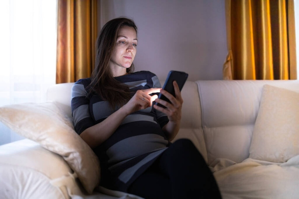Vrouwelijk browsen haar smartphone in haar gezellige appartement tijdens de avond - Foto, afbeelding