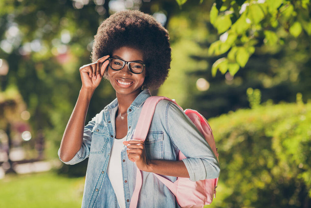 Photo portrait of pretty black skinned curly girl smiling happily wearing denim casual closes touching eyewear keeping pink bag - Photo, image