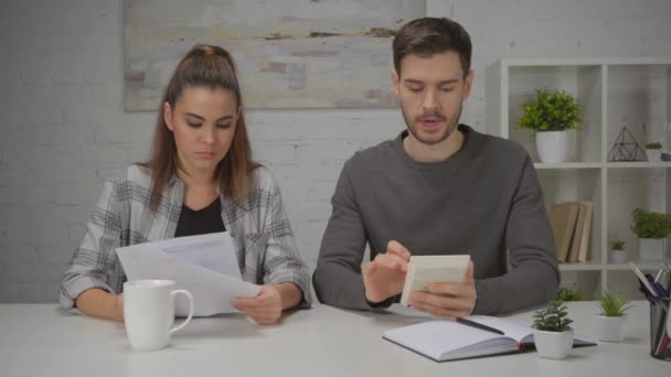 niezadowolona kobieta trzyma papiery i patrzy na mężczyznę używając kalkulatora - Materiał filmowy, wideo