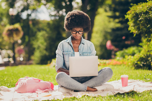 Photo portrait of black skinned young female student studying doing project on laptop sitting in park wearing glasses near bag cup of coffee - Foto, Bild