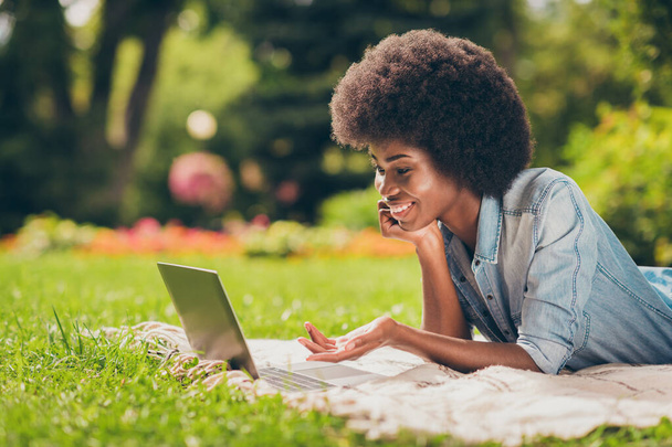 Side profile photo of black skinned young female student lying on grass having web video call smiling gesturing wearing jeans clothes - Fotó, kép