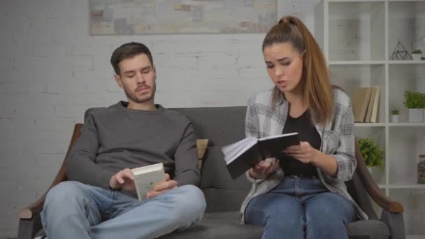 niezadowolona kobieta trzyma notatnik i kłóci się z mężczyzną siedząc na kanapie - Materiał filmowy, wideo