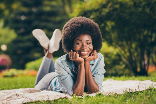Portrait photo of happy smiling black skinned girl laying on the grass in city park keeping hands near face wearing denim clothes - Fotó, kép