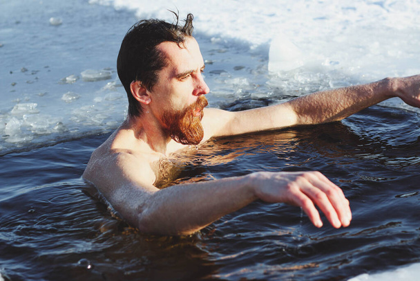 young, slim, handsome, sporty man with a red beard and long hair, naked, in black shorts, diving into ice-cold water in winter, against the backdrop Ukraine, Shostka - Фото, зображення