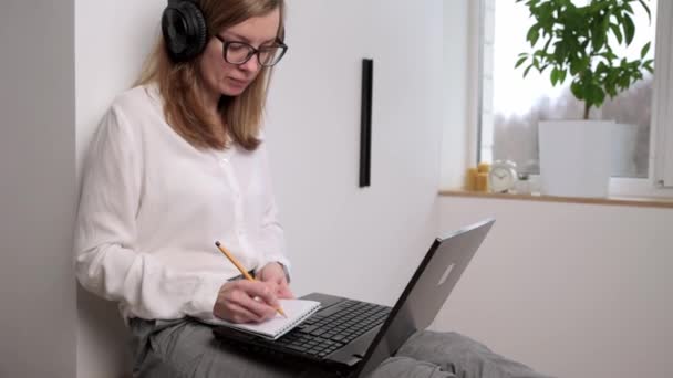 遠隔作業だ。ヘッドフォンの女性はノートブックでノートを作り、ノートパソコンのキーボードで印刷します。距離教育とeラーニングの概念 - 映像、動画