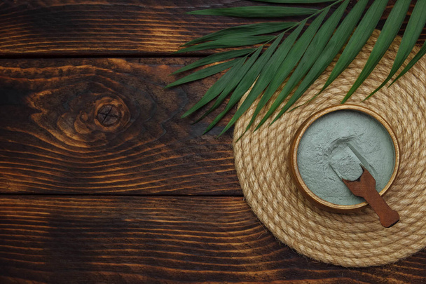 Dry Cosmetic clay in a bowl with a spoon on a wicker napkin with a green palm leaf on a brown wooden burnt background top view with a blank space for text, spa and skin care concept, template - Valokuva, kuva