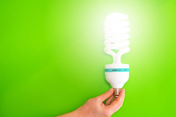 Close-up de uma mão segurando uma lâmpada led acesa contra um fundo verde. Conceito de economia de energia em casa. Espaço de cópia - Foto, Imagem