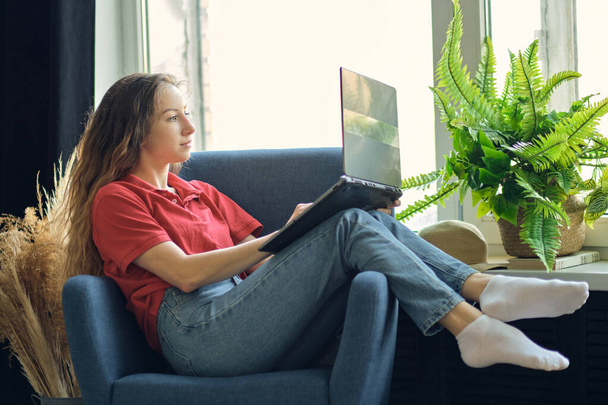 Young female using laptop after work, communicates on Internet with customer. Happy freelance woman is relaxing on comfortable couch and using laptop at home. - Valokuva, kuva