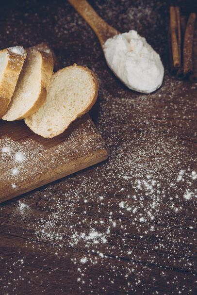 Flour in a wooden spoon, sliced French baguette on a wooden board, and cinnamon sticks sprinkled with flour on a wooden table. side view with place for text or logo. Retro photo. - Photo, image