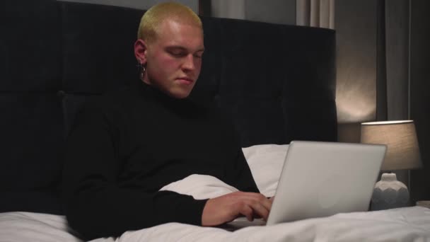 Hombre homosexual caucásico serio trabajando y escribiendo en el portátil en la cama tarde en la noche antes de quedarse dormido. - Metraje, vídeo