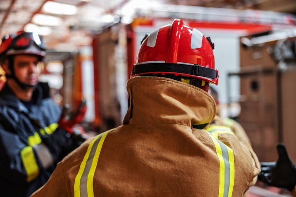 Rückansicht eines Feuerwehrmannes, der seine Schutzkleidung und seinen Helm anzieht, während er in der Feuerwehr steht. - Foto, Bild