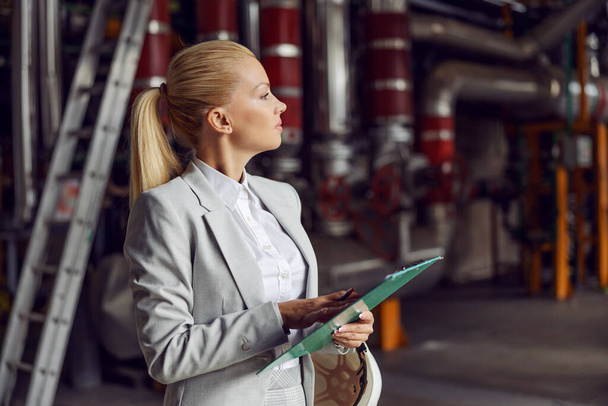 Blond businesswoman standing in heating plant, holding clipboard and checking on machinery. - Foto, Imagen