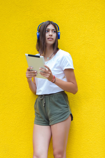 Junge Frau hört Musik auf digitalem Tablet über Kopfhörer an gelber Wand - Foto, Bild