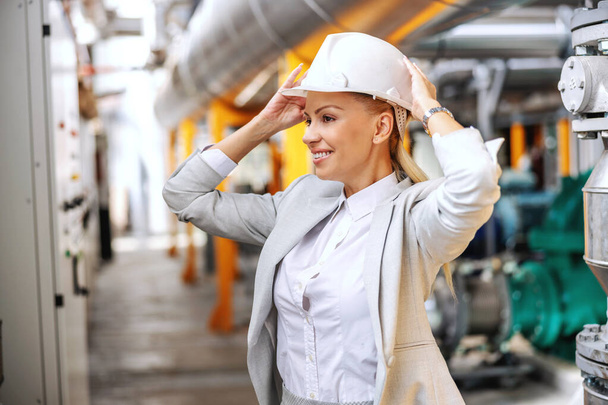 Lächelnde blonde Geschäftsfrau in offizieller Kleidung setzt Schutzhelm auf und bereitet sich auf den Gang durch das Kraftwerk vor. - Foto, Bild