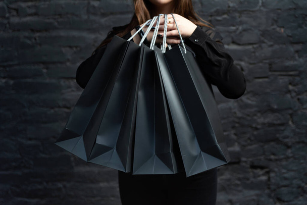 Μαύρο χαρτί τσάντες ψώνια σε γυναικεία χέρια σε μαύρο φόντο. Έννοια μαύρης Παρασκευής. - Φωτογραφία, εικόνα