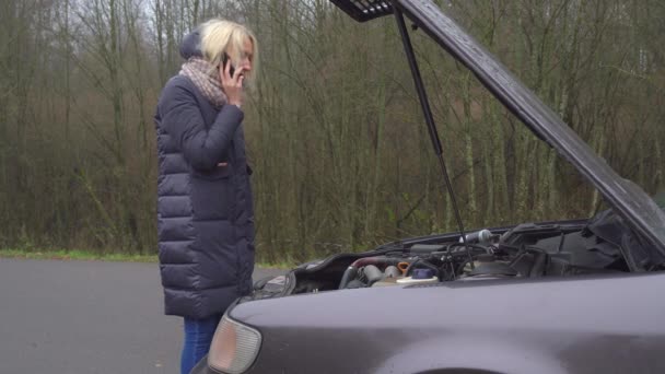 Egy nő telefonál az okostelefonján, miközben egy lerobbant autó mellett áll.. - Felvétel, videó