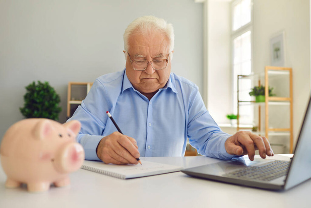 Retired man using laptop and taking notes in notebook studying pension plan and managing his budget - Zdjęcie, obraz