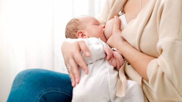 Młoda, opiekuńcza matka, siedząca na łóżku i karmiąca noworodka mlekiem. Pojęcie zdrowego i naturalnego żywienia piersią dla niemowląt. - Zdjęcie, obraz