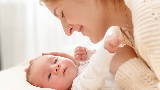 Zijaanzicht portret van blije lachende moeder strelen en knuffelen haar pasgeboren baby liggend op bed. Begrip gezinsgeluk en liefhebbende ouders met kleine kinderen - Foto, afbeelding
