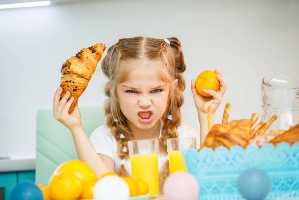A little girl of seven, wearing a white T-shirt, sits at the kitchen table. Holds ripe mandarine and a freshly baked croissant with chocolate - Foto, Bild