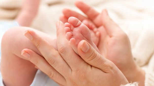 Closeup shot of young mother gently holding and touching little feet of her newborn baby boy lying in bed. Concept of family happiness and loving parents with little children - Photo, Image
