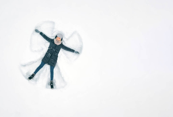 Joven hermosa mujer en una chaqueta gris, sombrero y guantes, pantalones vaqueros azules, botas hace un ángel de la nieve en la nieve, una vista desde un dron. Copiar espacio, pancarta. Nieve diversión, entretenimiento - Foto, imagen
