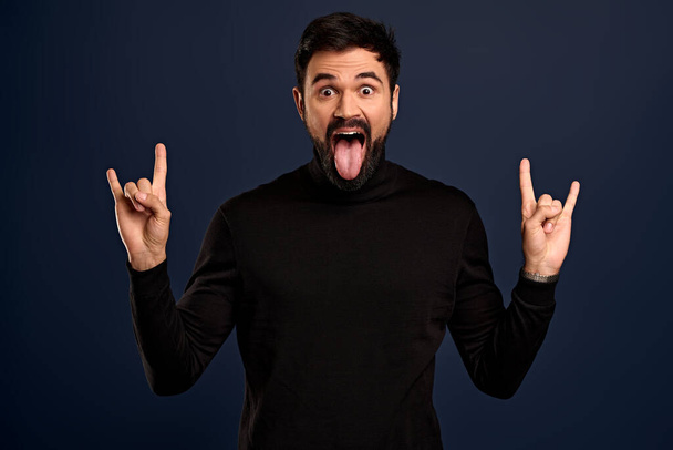 Fun, amusement and rock-n-roll concept. Good-looking overjoying modern guy enjoy music festival, show heavy metal gesture and tongue, squinting sassy, carefree feeling at concert, blue background. - Foto, immagini