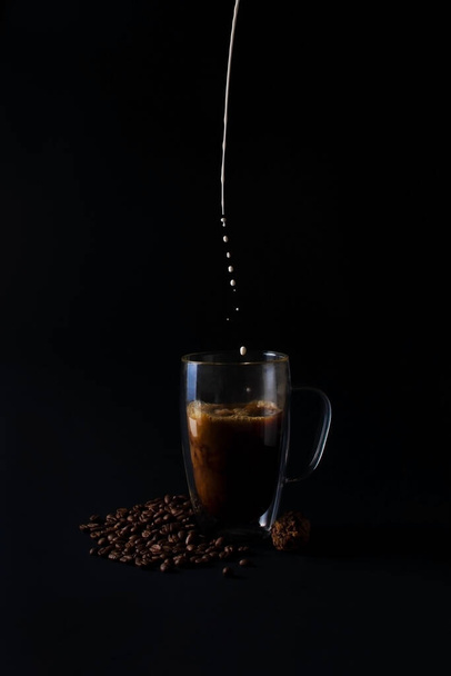 Milk is pouring into black coffee in glass cup on old dark background with beans and chocolate truffle. Breakfast coffee love concept. Front view - Фото, зображення
