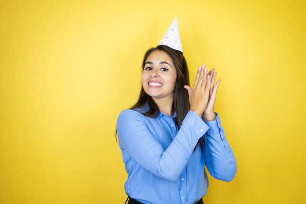 Young caucasian woman wearing a birthday hat over isolated yellow background clapping and applauding happy and joyful, smiling proud hands together - Photo, Image