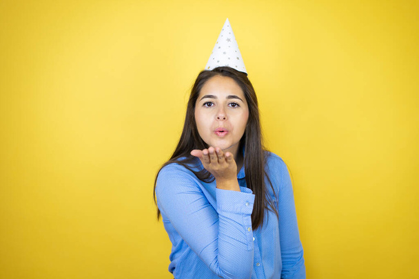 Young caucasian woman wearing a birthday hat over isolated yellow background looking at the camera blowing a kiss with hand on air being lovely and sexy. Love expression. - Photo, Image