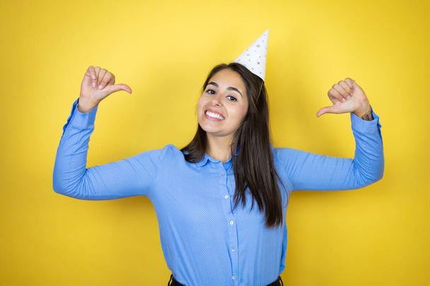 Jonge blanke vrouw draagt een verjaardagshoed over geïsoleerde gele achtergrond die er zelfverzekerd uitziet met een glimlach op het gezicht, wijzend met de vingers trots en gelukkig. - Foto, afbeelding