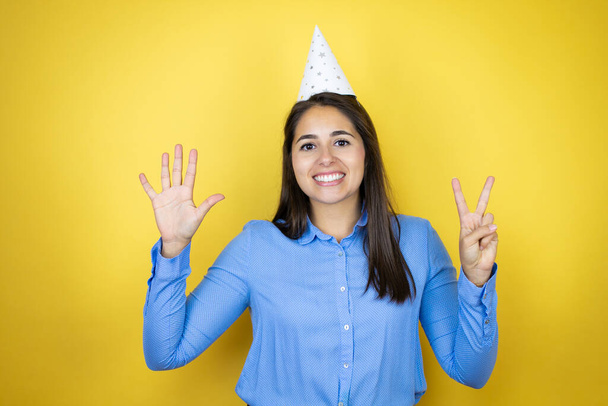 Young caucasian woman wearing a birthday hat over isolated yellow background showing and pointing up with fingers number seven while smiling confident and happy - Foto, Imagem