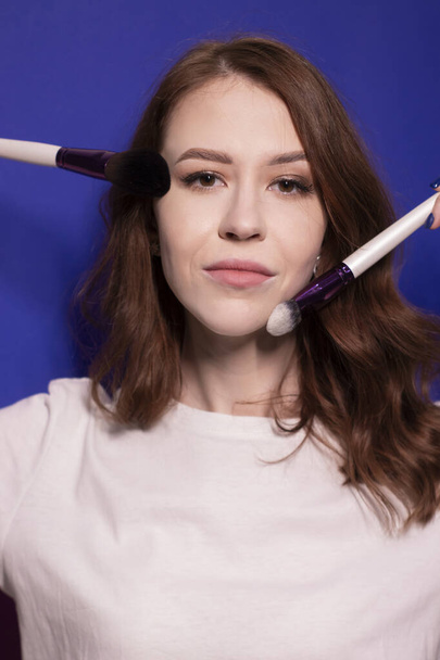 okouzlující brunetka žena v bílém tričku s make-up kartáčky izolované na barevném modrém pozadí. profesionální maskérka. kopírovat prostor - Fotografie, Obrázek