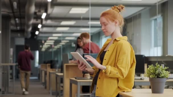 Keskipitkä hidasliikkeinen kuva nuoresta punatukkaisesta naispuolisesta toimistotyöntekijästä kirkkaan keltaisessa paidassa, joka työskentelee tabletin päällä seisoen isossa tilavassa toimistossa, kun taas työtoverit kommunikoivat taustalla - Materiaali, video