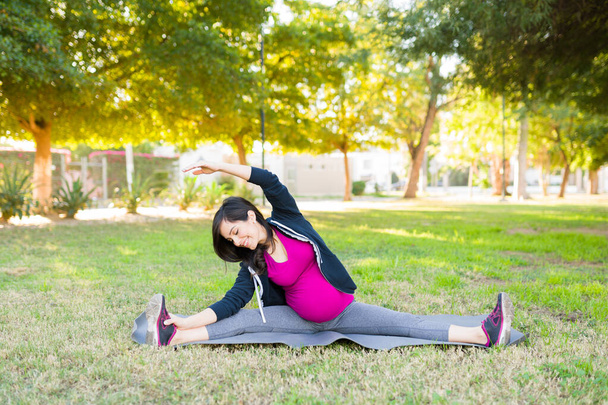 Mujer embarazada sonriente disfrutando de su entrenamiento en el parque durante un día soleado. Mujer feliz estirando sus brazos y piernas en una esterilla de yoga - Foto, Imagen