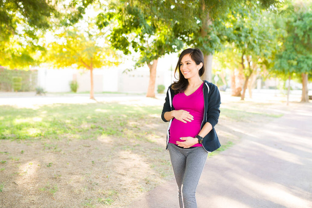 Femme enceinte rayonnante en vêtements de sport faisant une promenade dans le parc pendant une journée ensoleillée. femme heureuse profiter de ses exercices d'entraînement à l'extérieur - Photo, image