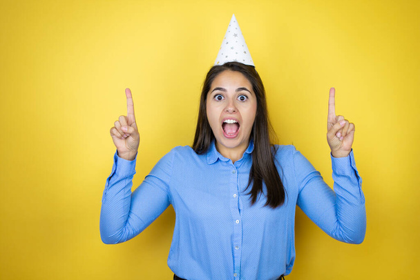 Young caucasian woman wearing a birthday hat over isolated yellow background amazed and surprised looking at the camera and pointing up with fingers and raised arms - Foto, immagini