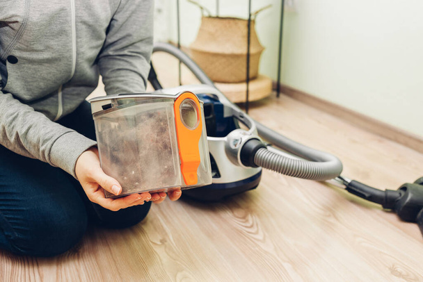 Mujer sosteniendo el filtro de polvo de la aspiradora en casa sentado en el suelo. Envase Hoover lleno de suciedad y basura. Funciones domésticas - Foto, Imagen