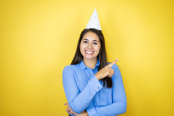 Mulher caucasiana jovem usando um chapéu de aniversário sobre fundo amarelo isolado sorrindo e apontando com a mão e o dedo para o lado - Foto, Imagem