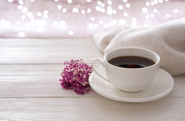 Eine weiße Tasse Kaffee, ein Strauß und ein Pullover vor dem Hintergrund einer brennenden Girlande. - Foto, Bild